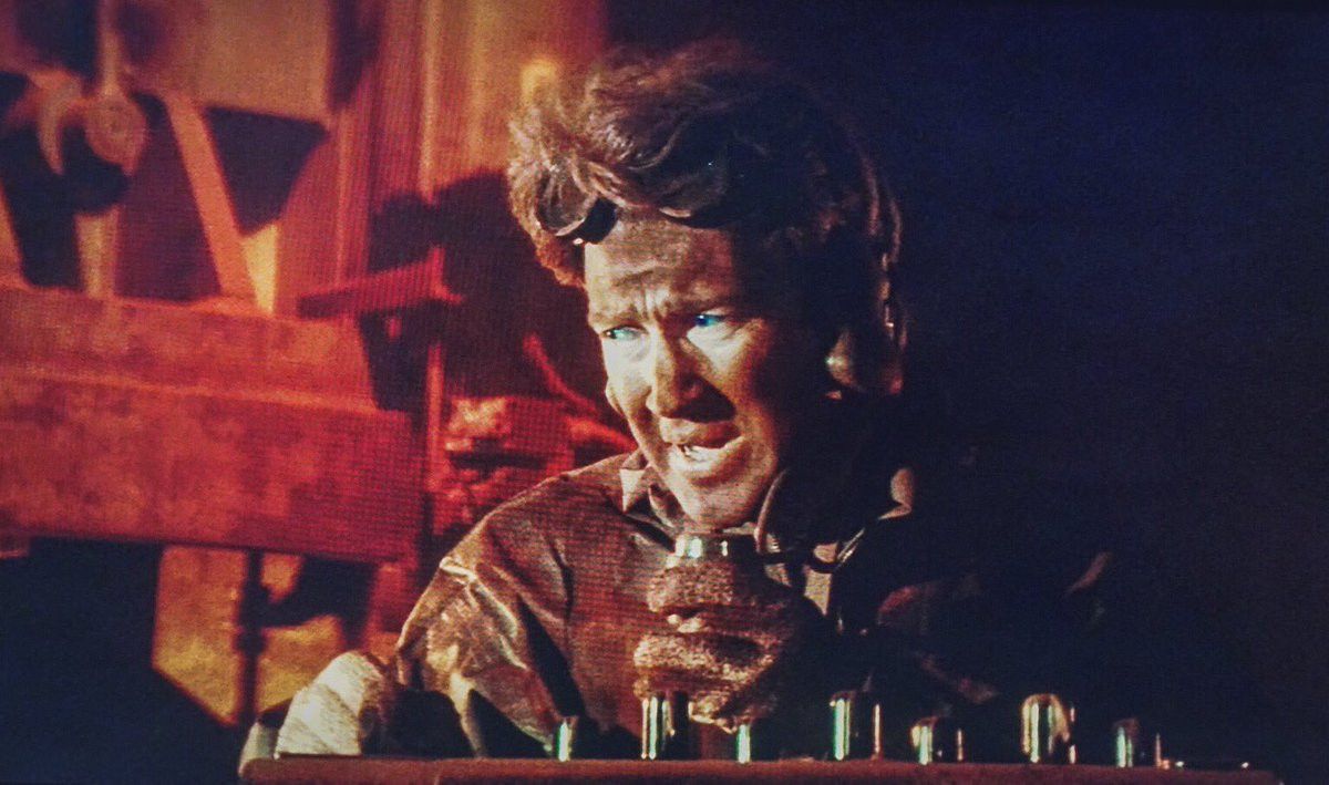 Screen grab from David Lynch's Dune. A Mid closeup shot of David Lynch's cameo appearance as the captain of a Spice Crawler. He is lit in half light and half shadow speaking into a microphone. Goggles on his head. 