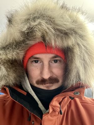 Selfie of a mustached man looking into the camera wearing a large parka with a faux fur hood. A hooded sweatshirt is visible under the turned up hood of the jacket. He is wearing a neck gaiter, and an orange winter had on underneath the hood. A slight smile visible.