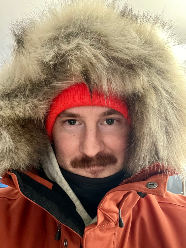 Selfie of a mustached man looking into the camera wearing a large parka with a faux fur hood. A hooded sweatshirt is visible under the turned up hood of the jacket. He is wearing a neck gaiter, and an orange winter had on underneath the hood. A slight smile visible.