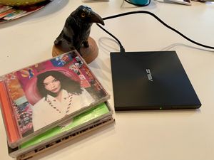 Picture of an external CD drive and a stack of compact discs next to it. A figurine of a crow is sitting near the stack of CDs watching over the procedure.