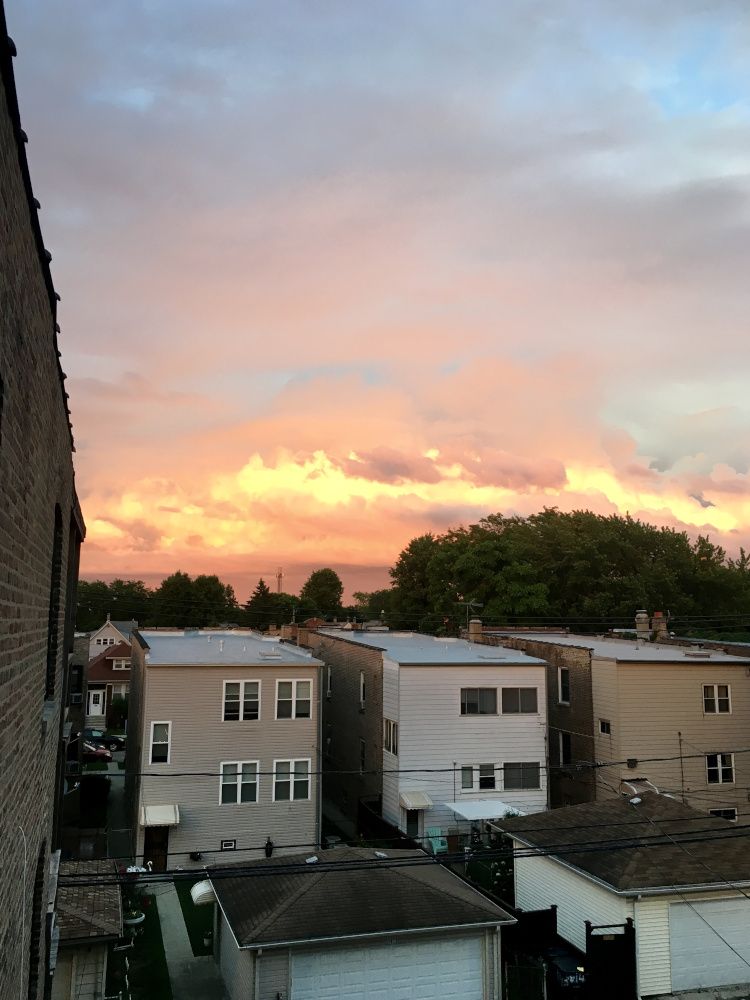 View from the third floor deck of a Chicago walk up looking East across an alleyway during a sunset. In the foreground is part of the building the photo is being taken. Midground has the alleyway, alley-facing garages, and the houses on the other side of the block. In the distance are clouds blazing orange with the light from the setting sun.