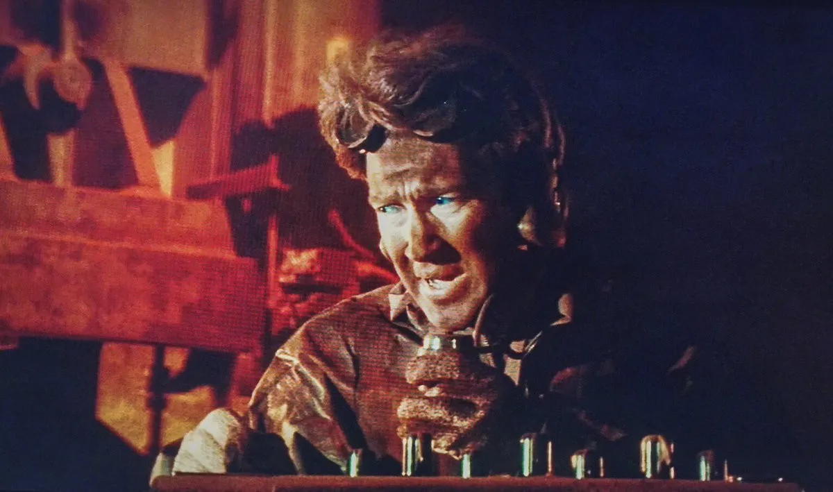 Screen grab from David Lynch's Dune. A Mid closeup shot of David Lynch's cameo appearance as the captain of a Spice Crawler. He is lit in half light and half shadow speaking into a microphone. Goggles on his head.
