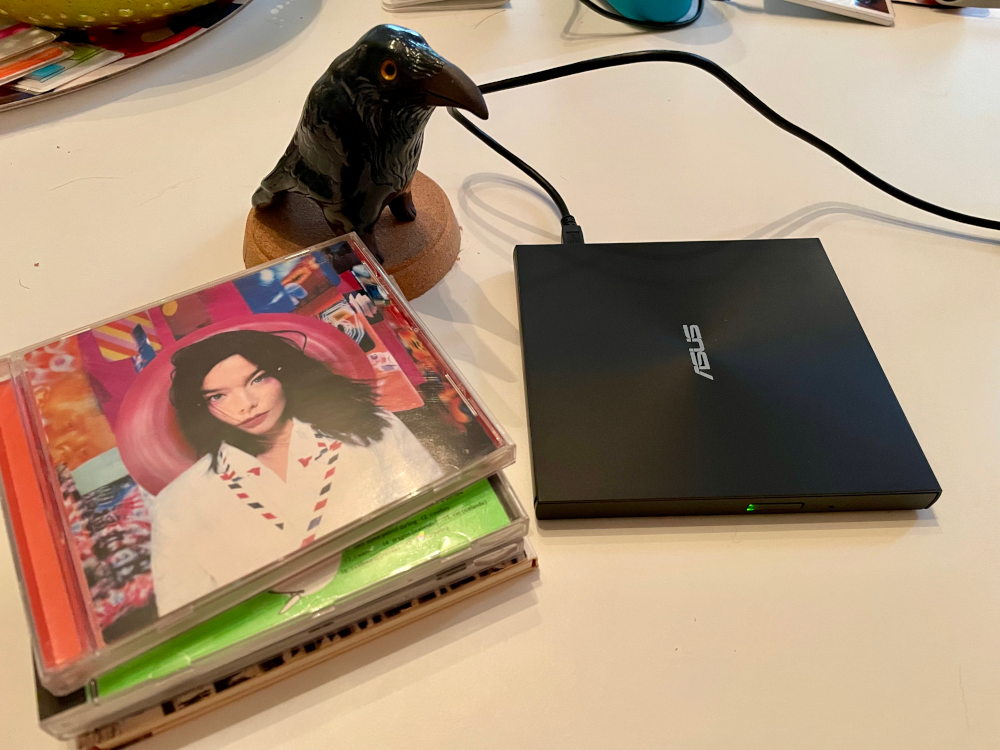 Picture of an external CD drive and a stack of compact discs next to it. A figurine of a crow is sitting near the stack of CDs watching over the procedure.